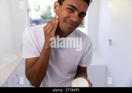 Glücklicher birassischer Mann, der zu Hause im Badezimmer Gesichtscreme aufträgt Stockfoto