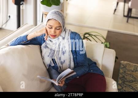 Birassische Frau im Hijab, die Buch im Wohnzimmer liest Stockfoto