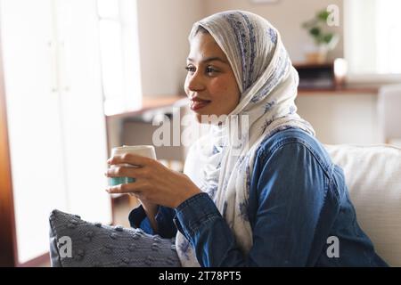 Glückliche birassische Frau im Hijab, die Kaffee im Wohnzimmer trinkt Stockfoto