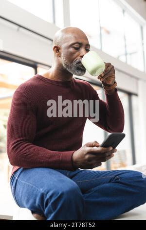 Reifer afroamerikanischer Mann trinkt Kaffee mit Smartphone, der in der Küche sitzt Stockfoto