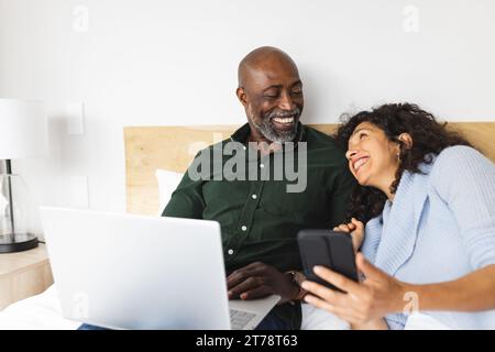 Glückliches reiches Ehepaar, das im Schlafzimmer mit Laptop und Smartphone auf dem Bett sitzt Stockfoto