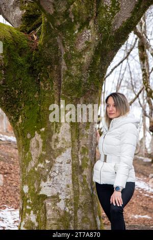 Eine Frau in einer weißen Jacke steht neben einem moosbedeckten Baum im Wald Stockfoto