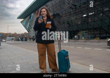 Geschäftsfrau, die sich vom Flughafen beeilt, auf der Straße der Stadt steht und einen Pass mit Tickets hält und einen Koffer bei sich trägt, während sie ein Taxi per Handy anruft Stockfoto