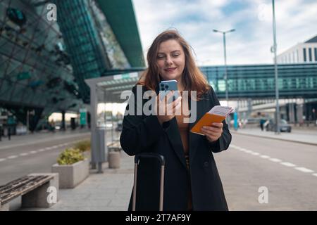 Geschäftsfrau, die sich vom Flughafen beeilt, auf der Straße der Stadt steht und einen Pass mit Tickets hält und einen Koffer bei sich trägt, während sie ein Taxi per Handy anruft Stockfoto