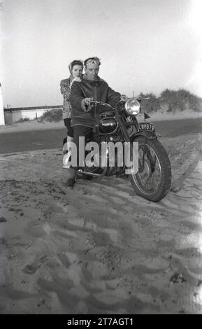 1950er Jahre, historisch, draußen, an einem Sandstrand, ein Mann in einer ledernen Bomberjacke, Armeetyp und Gesichtsbrille und eine Frau, die einen Schal trägt, sitzt zusammen auf einem Triumph Motorrad der Ära, England, Großbritannien. Das britische Motorrad wurde in Coventry von der Triumph Engineering Co hergestellt, deren Geschichte bis ins Jahr 1883 zurückreicht, und Siegfried Bettman, ein Immigrant aus Deutschland, der ein Import-/Exportgeschäft für Fahrräder gründete. Stockfoto