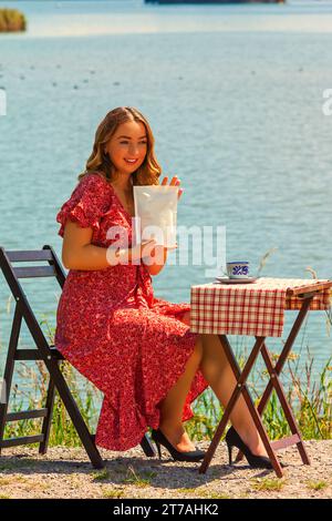 Frau, die im Sommer am See sitzt und ein Kaffeepaket in der Hand hält Stockfoto