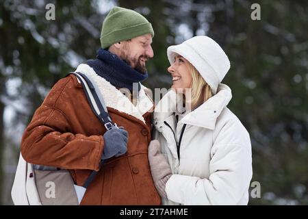 Glückliches junges, liebevolles Paar in Winterkleidung, das sich im Winterwald vor der Kamera ansieht Stockfoto