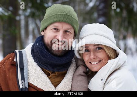 Liebevoll lächelndes Paar in Winterkleidung, das vor der Kamera steht und Sie im Wald mit immergrünen, schneebedeckten Bäumen ansieht Stockfoto