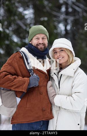 Junges, liebevolles Paar in warmen Wintermützen und Mänteln, die in die Kamera schauen, während sie im Winterwald stehen und Urlaub genießen Stockfoto