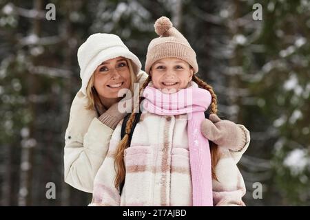 Glückliche junge, liebevolle Mutter, die ihre Tochter in Winterkleidung umschließt, während sie im Winterwald steht und in die Kamera blickt Stockfoto