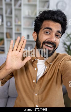 Glücklicher Hindu-Mann, der per Videoanruf am Telefon spricht, Begrüßung winkt zur Kamera, plaudert mit Familie und Freunden, lächelt, Kameraansicht vertikal. Stockfoto