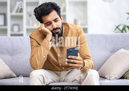 Trauriger hindu-Mann, der zu Hause in einem hellen Zimmer auf dem Sofa sitzt, das Telefon in der Hand hält und auf den Bildschirm blickt, traurig wegen schlechter Nachrichten, gelangweilt, Kopf mit der Hand hält. Stockfoto