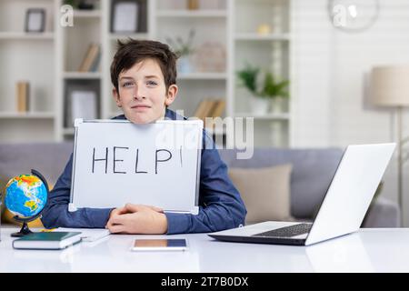 Trauriger müder Teenager, der an einem Tisch vor einem Laptop sitzt und ein Schild mit dem Wort Hilfe in der Hand hält, Bildungskonzept, Lernschwierigkeiten. Stockfoto