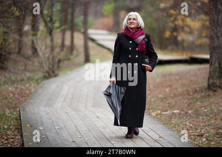 Schöne Seniorin mit Regenschirm spaziert entlang des Waldes, trägt stilvolle trendige Kleidung, einen warmen langen Mantel. Stockfoto