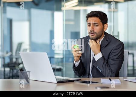 Unglücklicher hinduistischer Geschäftsmann, der an Halsschmerzen leidet, mit Kehlspray in der Hand hält, im modernen Büro sitzt und Hand an Kehle hält. Stockfoto