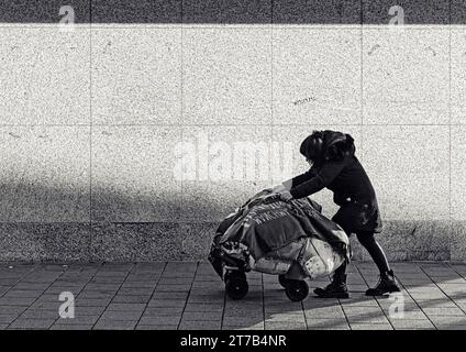 Obdachlose Frau, die eine Straße entlang läuft und ihre Sachen auf einem Trolley drückt Stockfoto