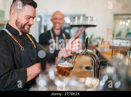 Stilvoller bärtiger Barkeeper in schwarzer Uniform, der Bier an der Bar anzapft und der Kellner mit Tablett plauderte miteinander und lächelte miteinander. Erfolgreiches People Team Wo Stockfoto