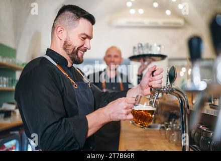 Stilvoller bärtiger Barkeeper in schwarzer Uniform, der Bier an der Bar anzapft und der Kellner mit Tablett plauderte miteinander und lächelte miteinander. Erfolgreiches People Team Wo Stockfoto