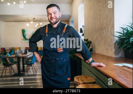 Porträt eines glücklichen, lächelnden kleinen Geschäftsinhabers in schwarzer Küchenuniform mit Schürze in seinem gemütlichen Restaurantsaal. Erfolgreiche Menschen, harte Arbeit Stockfoto
