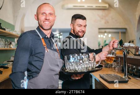Stilvoller bärtiger Barkeeper in schwarzer Uniform, der Bier an der Bar anzapft und der Kellner mit Tablett plauderte miteinander und lächelte miteinander. Erfolgreiches People Team Wo Stockfoto
