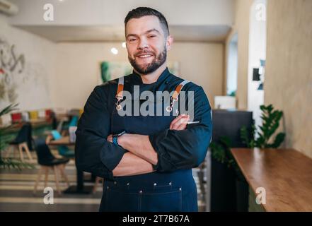 Porträt eines glücklichen, lächelnden kleinen Geschäftsinhabers in schwarzer Küchenuniform mit Schürze in seinem gemütlichen Restaurantsaal. Erfolgreiche Menschen, harte Arbeit Stockfoto