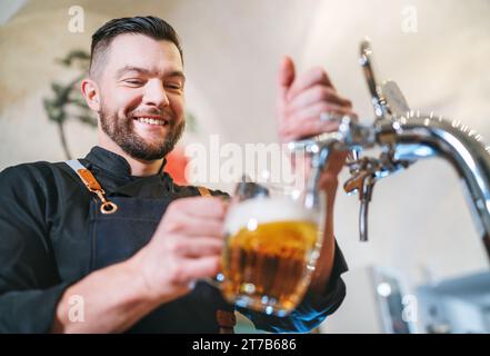 Ein lächelnder, stilvoller Bärenbarmann in schwarzer Uniform mit einer Schürze, der frisches Lagerbier in einen Pilsner-Glas-Becher an der Bar zapft. Erfolgreiche Mitarbeiter, Stockfoto