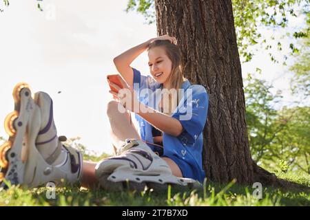 Frau, die im Park Schlittschuhlaufen kann. Mädchen, die auf dem Gras sitzen und sich mit Inline-Skates entspannen, während sie sich auf dem Smartphone unterhalten. Kaukasische Frau bei Outdoor-Aktivitäten. Stockfoto