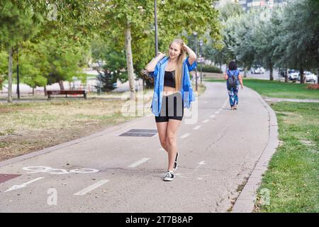 Die junge weiße Frau mit blonden Haaren geht mit Inline-Rollschuhen in der Hand in einen Park, um zum Skating-Training zu gehen und Sport auf einem heißen S zu treiben Stockfoto
