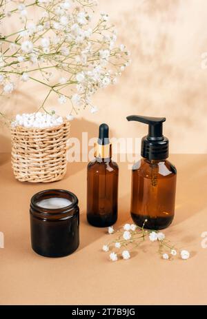 Kosmetischer Gesichtsreiniger, braune Glasflaschen des Serums, Cremebecher und weiße Blüten der gypsophila auf hellbeigem Hintergrund. Naturkosmetik, Spa und Wellness Stockfoto