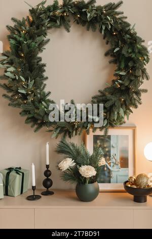 Beleuchteter Weihnachtskranz, der an einer Wand über einem Sideboard mit gewickelten Geschenken, Kerzen, Blumen, Lampe und Bilderrahmen hängt Stockfoto