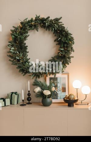 Beleuchteter Weihnachtskranz, der an einer Wand über einem Sideboard mit gewickelten Geschenken, Kerzen, Blumen, Lampe und Bilderrahmen hängt Stockfoto