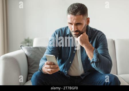 Ein nachdenklicher Mann mittleren Alters Doom blättert zu Hause durch das Telefon Stockfoto