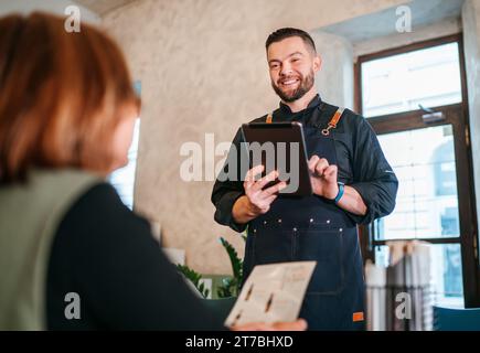 Lächelnder Kellner aus Kaukasier, der Bestellungen von weiblichen Kunden mit digitalem Tablet im Café-Restaurant entgegennimmt. Glückliche Menschen, Restaurants, Fastfood und Food-Service Stockfoto