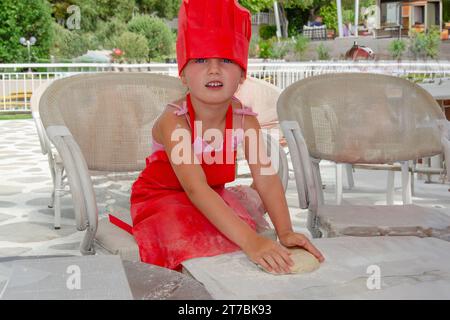 Ein fünfjähriges Mädchen in rotem Kochmütze und Schürze knetscht Teig. Sie ist auf der Sommerterrasse mit weißen Gartenstühlen und Tisch. Stockfoto