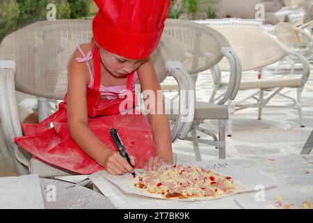 Ein fünfjähriges Mädchen in roter Küchenschürze macht Pizza. Sie ist auf der Sommerterrasse mit weißen Stühlen und Tisch Stockfoto
