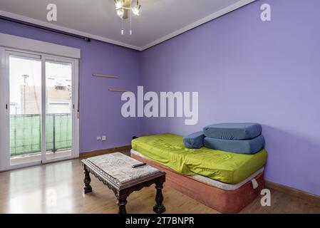 Ein kleines Wohnzimmer in einem Haus mit einem Couchtisch und einem unordentlichen Bett, Holzböden, Zugang zu einer Terrasse mit Aluminium- und weißen Glastüren und Violett Stockfoto