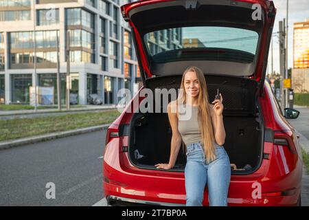 Junge blonde Frau, die im Kofferraum eines neuen roten Autos sitzt und Schlüssel hält. Stockfoto