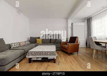 Ein geräumiges Wohnzimmer mit einem Ecksofa mit grauem Stoff, einem quadratischen Couchtisch mit Tischdecke, Stockfoto