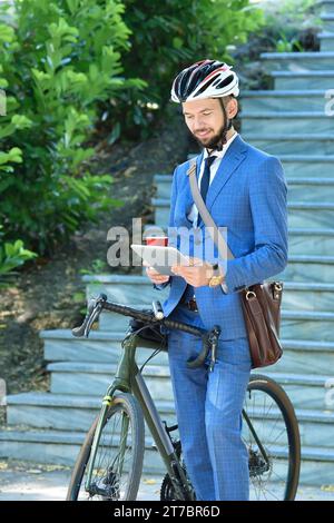 Bärtiger Geschäftsmann mit Fahrrad mit Kaffeetasse und digitalem Tablet. Business- und Alternativverkehrskonzept Stockfoto