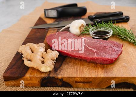 Rohes Rindersteak mit Ingwerwurzel, Knoblauch, Sojasauce und Rosmarin auf einem Holzbrett auf dem Küchentisch Stockfoto