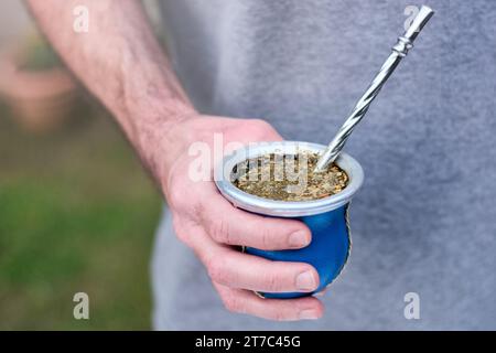 Hand eines jungen kaukasischen Mannes, der einen Mate-Becher hält, traditioneller Aufguss in vielen südamerikanischen Ländern. Unscharfer Hintergrund mit Kopierraum für Text Stockfoto