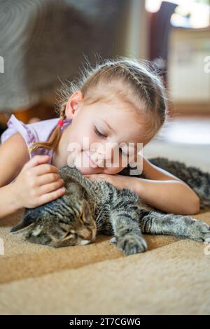 Kleines Mädchen streichelt graue Katze auf Teppich im Wohnzimmer zu Hause Stockfoto