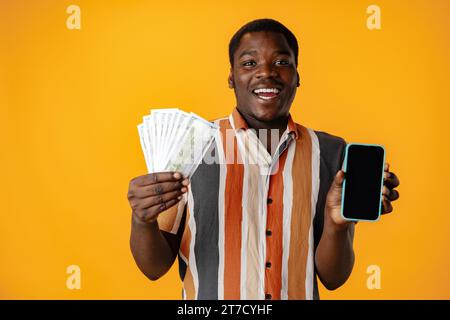 Junger afrikanischer Mann, der Smartphone- und Dollarscheine vor gelbem Hintergrund zeigt Stockfoto