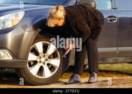 Unerkennbare blonde Frau lehnt sich über ihr Auto und wechselt an sonnigen Herbsttagen in der Natur kaputten Reifen. Tourismus und Reisen. Abenteuer und Urlaub Stockfoto