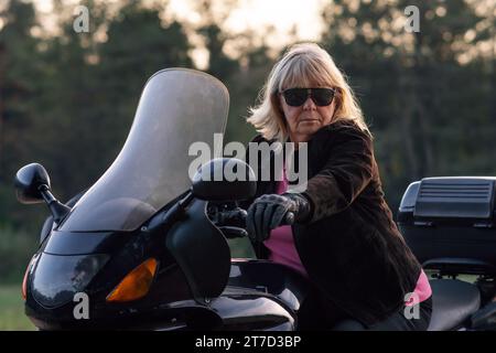 Stilvolle blonde Motorradtouristin, Supermutter, Großmutter in schwarzen Handschuhen und schwarzer Brille, sitzend auf ihrem Fahrrad. Hobby-Motorradtourismus. Gesundes li Stockfoto