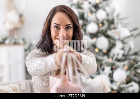 Gut aussehende Brünette händigt einen Haufen Papiertüten mit Geschenken in Richtung Kamera mit Weihnachtsbaum im Hintergrund. Stockfoto