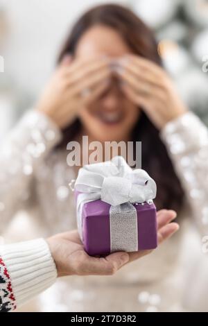 Die Hand des Mannes hält lila Weihnachtsgeschenk mit einem Band vor einer Frau, die ihre Augen mit den Händen schließt. Stockfoto