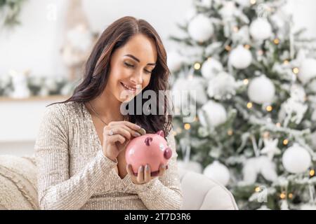 Eine lächelnde Frau steckt eine Münze in eine rosa Schweinebank, die neben einem Weihnachtsbaum sitzt. Stockfoto