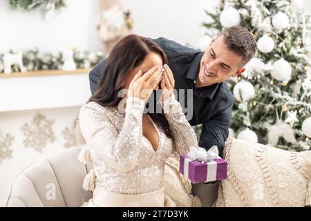 Der Ehemann hält ein lila kleines Weihnachtsgeschenk vor seiner Frau, während sie ihre Augen mit beiden Handflächen geschlossen hält. Stockfoto