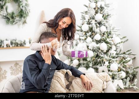 Die Frau hält ihre Hand vor die Hand des Mannes, als sie ihn mit einem Weihnachtsgeschenk überraschen will. Stockfoto